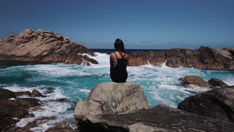 lonely and pensive girl sitting on the rock observes the rough ocean