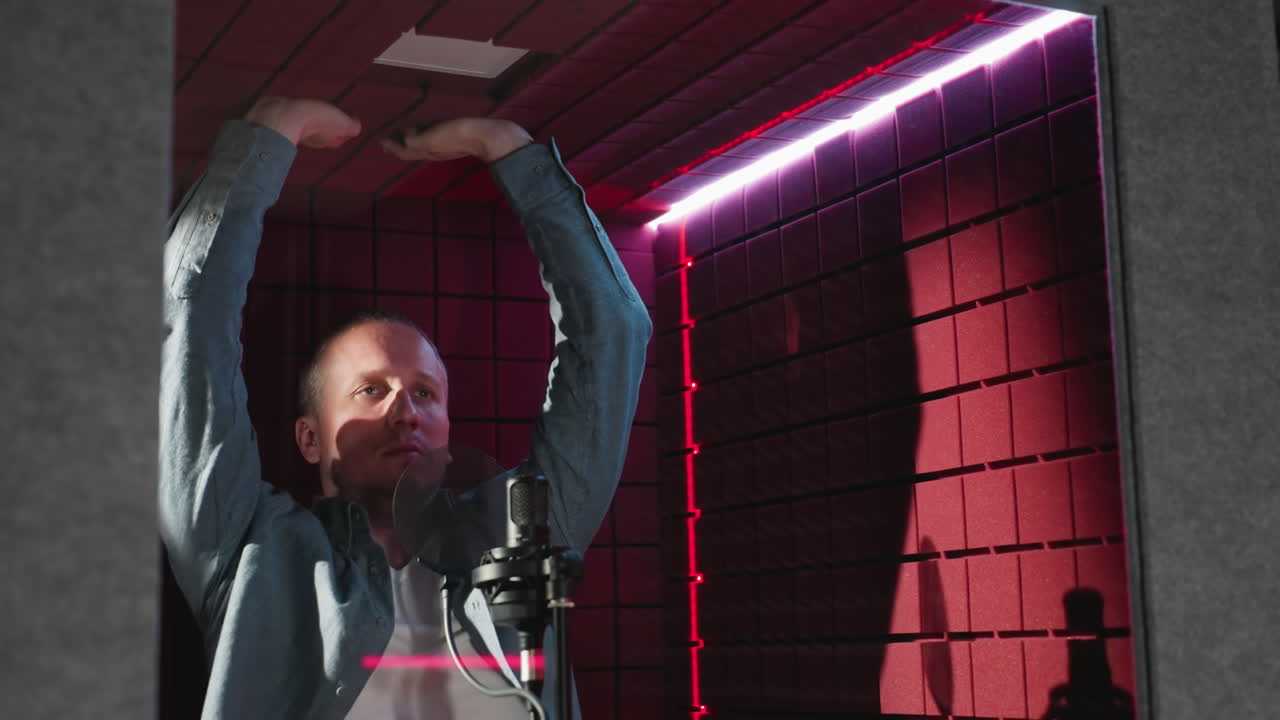 A man in a blue long-sleeve shirt and white inner stretches upwards, touching the ceiling and side walls of a red soundproof room, highlighted by vibrant LED lighting