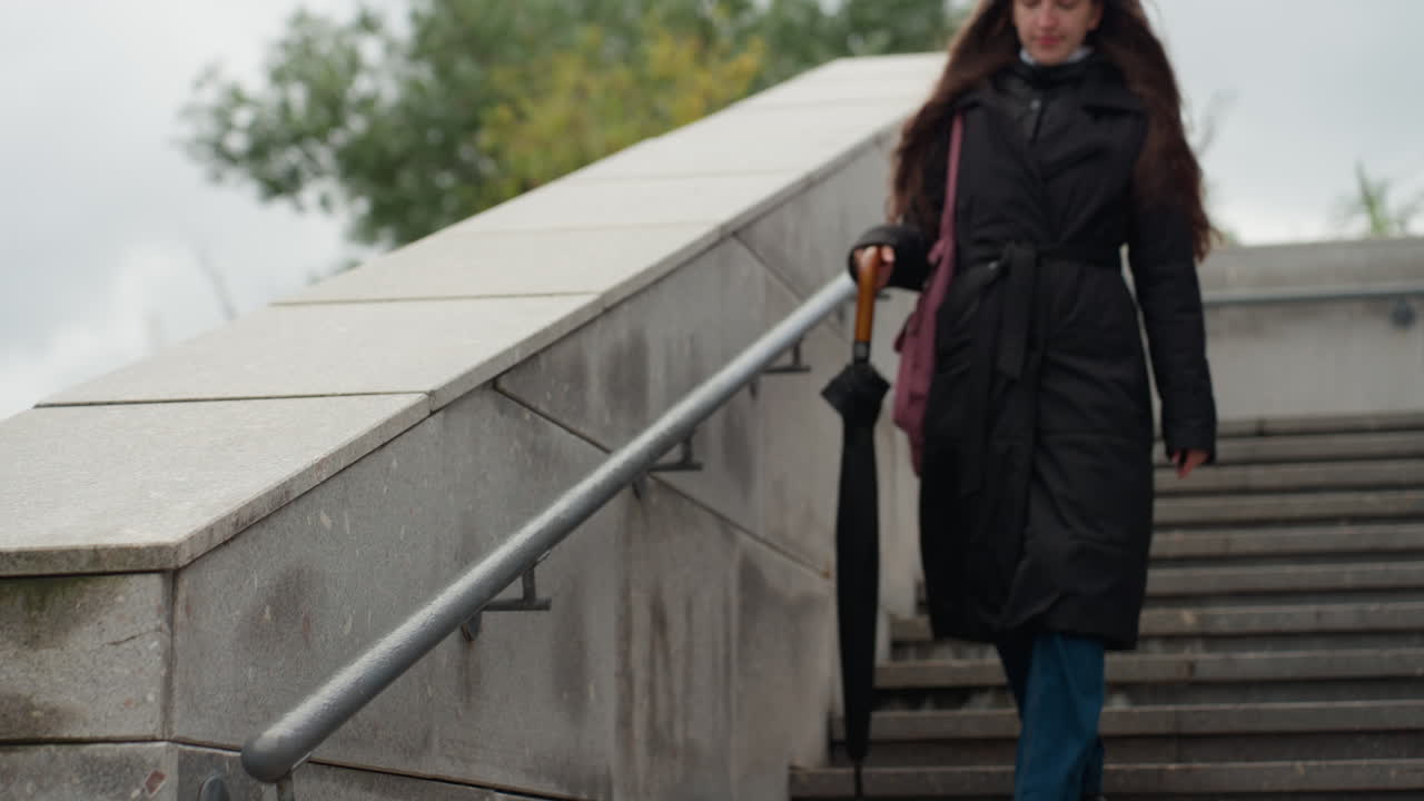 young woman walking down stairs wearing dark coat, carrying folded umbrella and shoulder bag, descending stone bridge steps under overcast sky, autumn trees and lamp post framing scene, long brunette