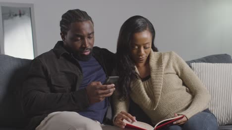 couple at home sitting on sofa in lounge with woman reading book and man looking at mobile phone