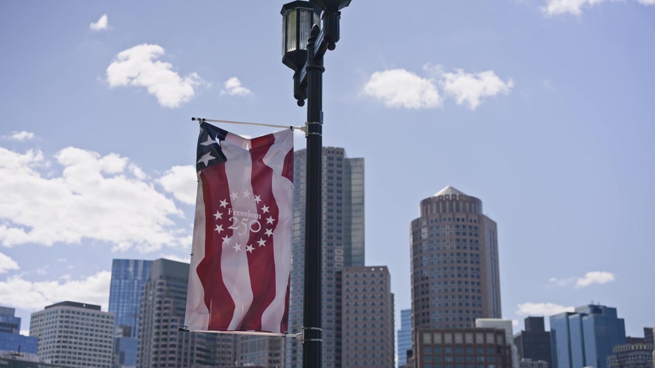 Carrellata cinematografica in avanti verso la bandiera americana che si affaccia sul porto di Boston e sullo skyline del centro di Boston. Sole splendente, viste tranquille sul lungomare e l'architettura iconica della città creano un'atmosfera.