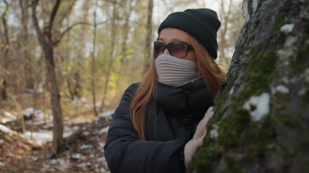 Mulher suspeita espreita por trás de uma bétula, examinando a trilha tranquila com a mão enluvada na casca, roupas de inverno em camadas e óculos de sol obscurecem a identidade, luz suave através dos galhos nus cria tensão