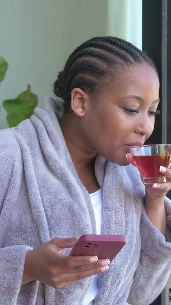Vídeo vertical: Mulher de roupão, lendo no celular rosa e tomando chá em uma xícara de vidro na varanda, relaxando.