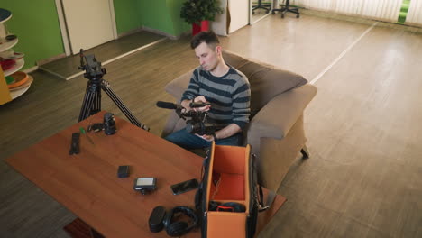 man sitting on couch reaching into camera equipment bag for gear. camera lenses, smartphone, microphone, and other video production tools on table. studio setting with green walls