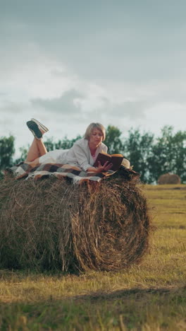 dama relajándose con la pierna levantada en una bola de heno con una manta a cuadros, leyendo un libro en un campo abierto, disfrutando de la serenidad de la naturaleza con la tranquila vista circundante del campo