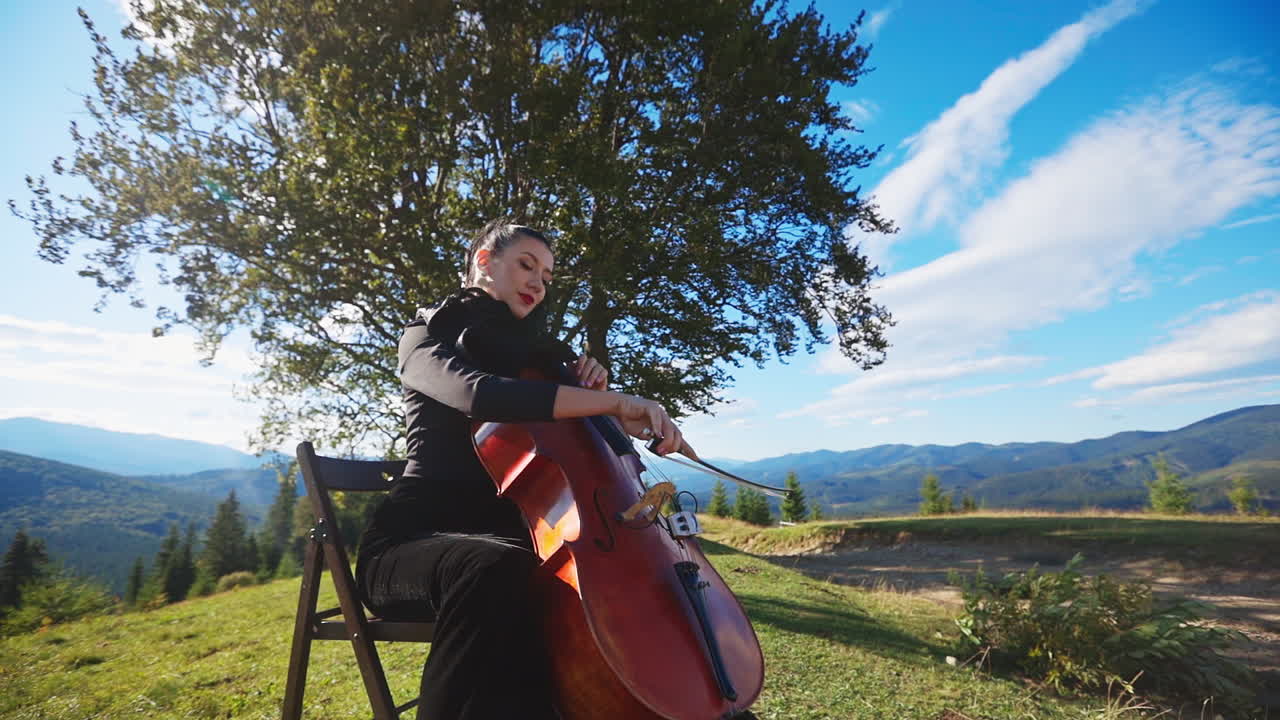 Solo music player sitting in the nature. Violoncello musician lady in black dress playing instrument in the beautiful mountainous area.