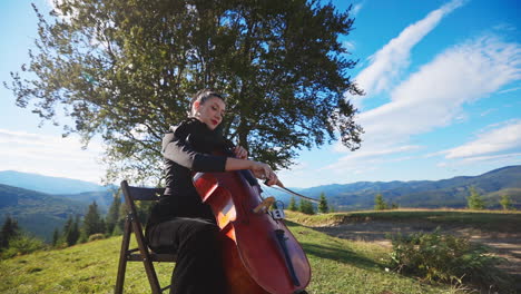 solo music player sitting in the nature. violoncello musician lady in black dress playing instrument in the beautiful mountainous area.