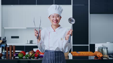 mujer chef asiática sonriendo y mostrando pinzas y cuchara a la cámara en la cocina de casa