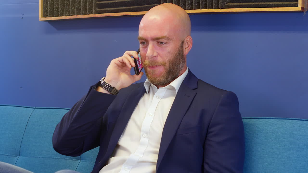 Uomo d'affari felice che parla e ride con un amico al telefono cellulare mentre è seduto sul divano nell'ufficio di casa