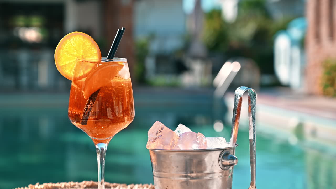 Cocktail and bucket with ice, slice of orange, ice tongs, pool on the background, Greece