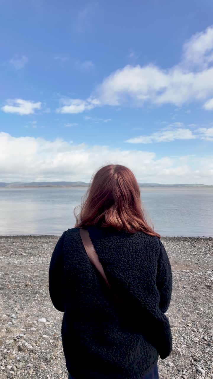 Kvinde praktiserer mindfulness og mentalt velvære, mens hun står på en stenstrand og kigger ud over en rolig havhorisont. Vertikal video.