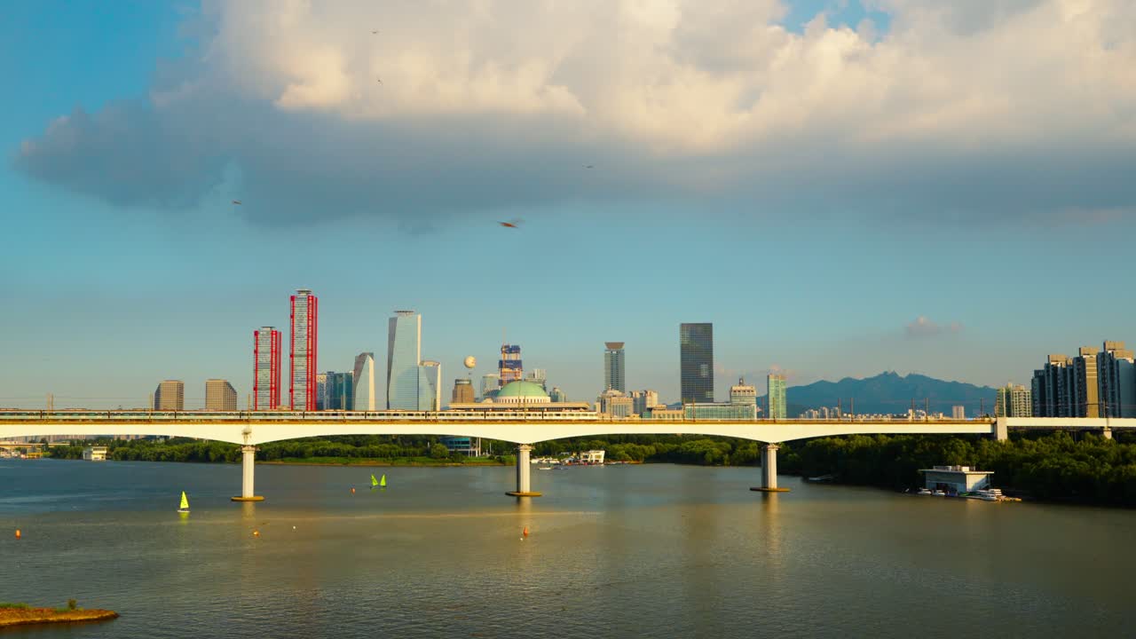 Seoul cityscape - subway train crossing Dangsan Railway Bridge on the Han River, with the Yeouido financial district skyline lit by golden sunset sunlight - static aerial view