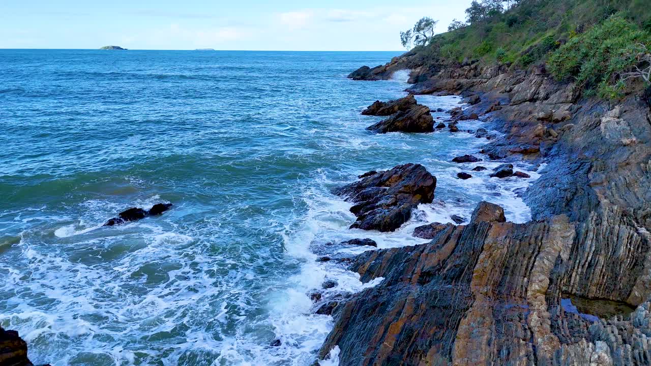 Dynamic ocean waves crash against rugged rocks under clear skies at Charlesworth Bay Beach, creating a vibrant coastal scene