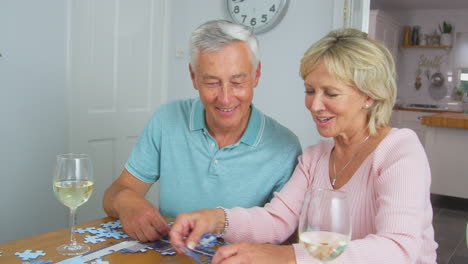 Älteres-Rentnerehepaar-Sitzt-Zu-Hause-Mit-Einem-Glas-Wein-Am-Tisch-Und-Macht-Puzzle