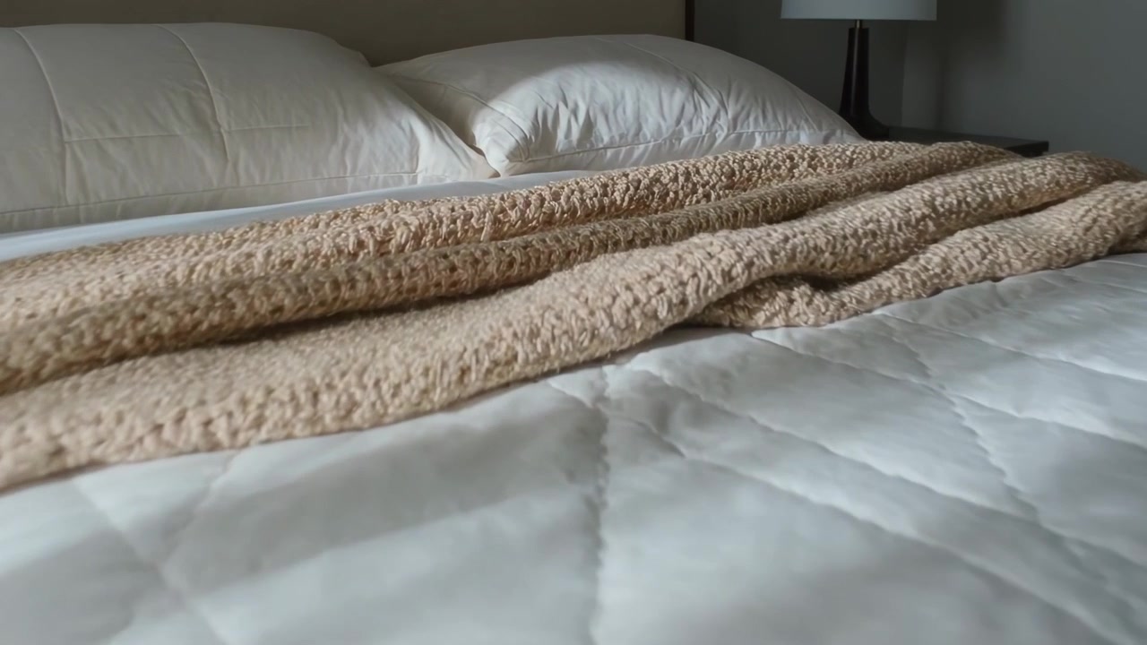 Telecamera in movimento che si concentra su un letto rifatto in camera da letto, mostrando la trama di una coperta in maglia beige, un piumino e cuscini.