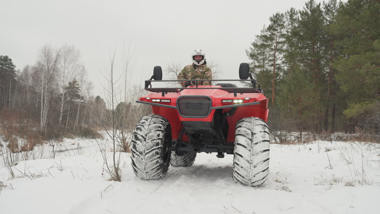ATV rosso al minimo su pista innevata, pilota che sistema il casco e scruta l'orizzonte, pneumatici robusti pieni di neve, bordo campo con alberi sparsi, cielo spento, composizione frontale stabile, ricognizione attenta.