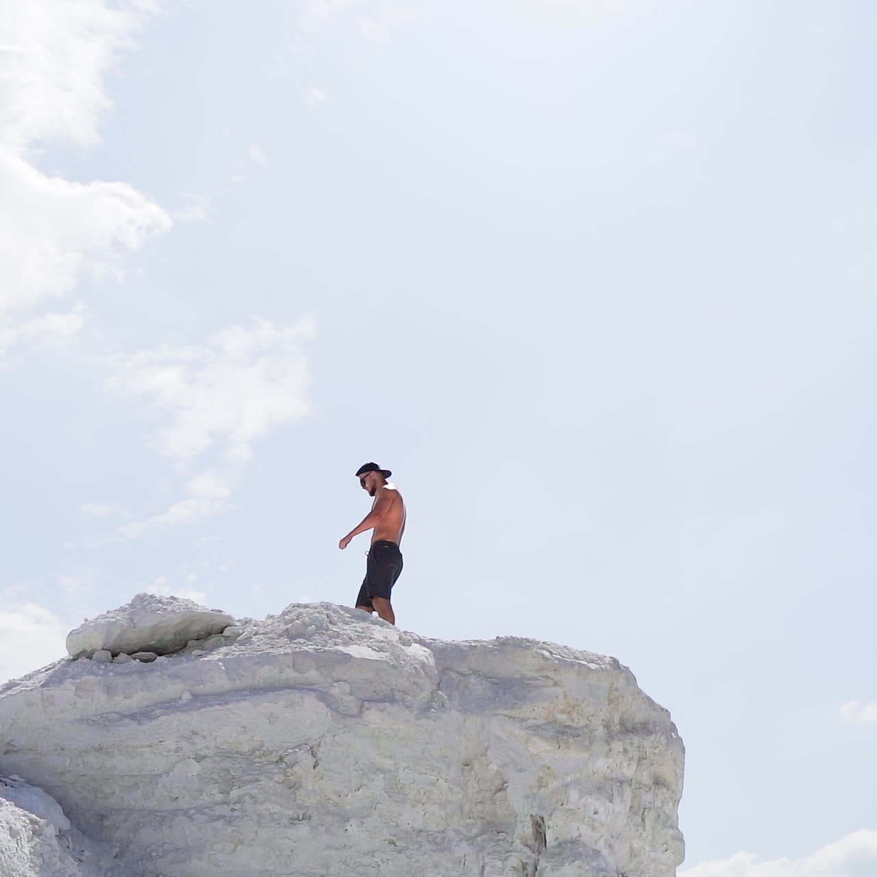 Sportive man on the edge of the hill. Shirtless man in shorts under the light sky. Muscular athlete on the top of mount in summer. View from below. Slow motion.