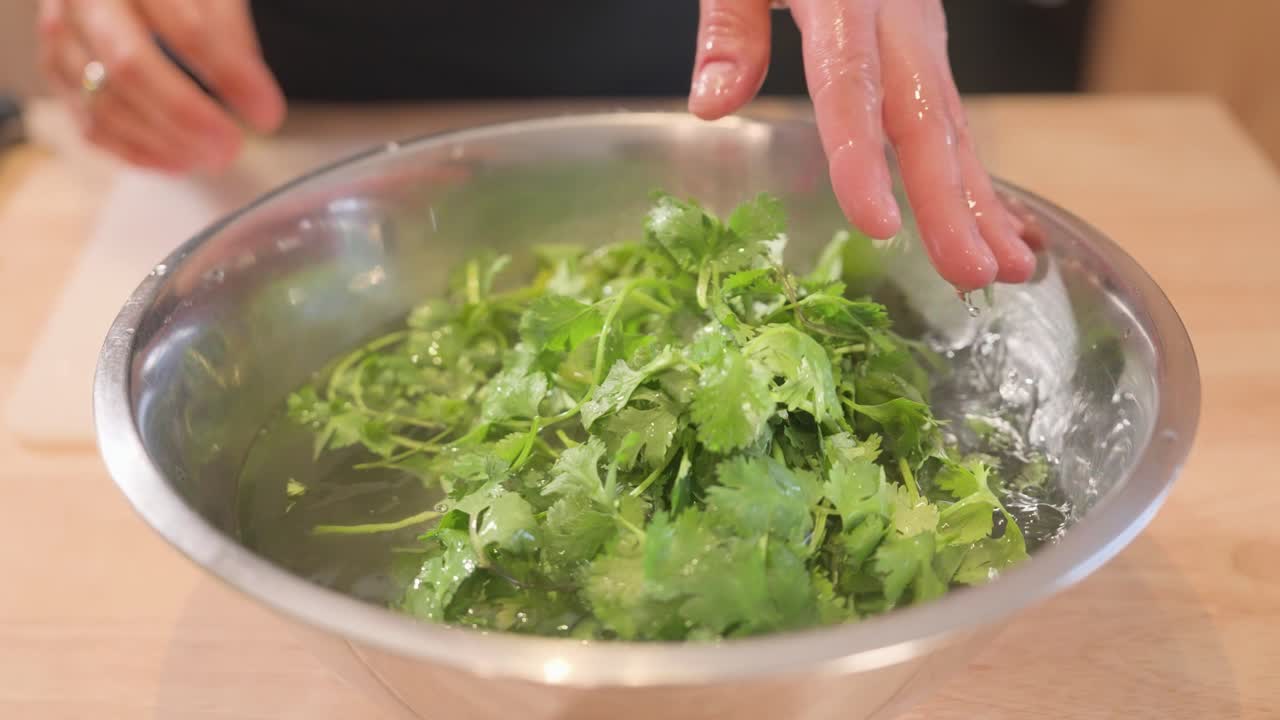 A hand rinses fresh cilantro in a stainless steel bowl filled with water on a wooden kitchen island.
