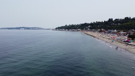 Drohnenaufnahme,-Die-Einen-Kontrast-Zwischen-Dem-Ruhigen-Wasser-Des-Puget-Sound-Und-Den-Belebten-Sandstränden-Von-Alki-Beach-In-Seattle-Bildet