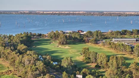 Antena-A-Través-Del-Campo-De-Golf-Hasta-Casas-Nuevas-Y-El-Lago-Mulwala-Y-árboles-Muertos-En-El-Agua-Más-Allá