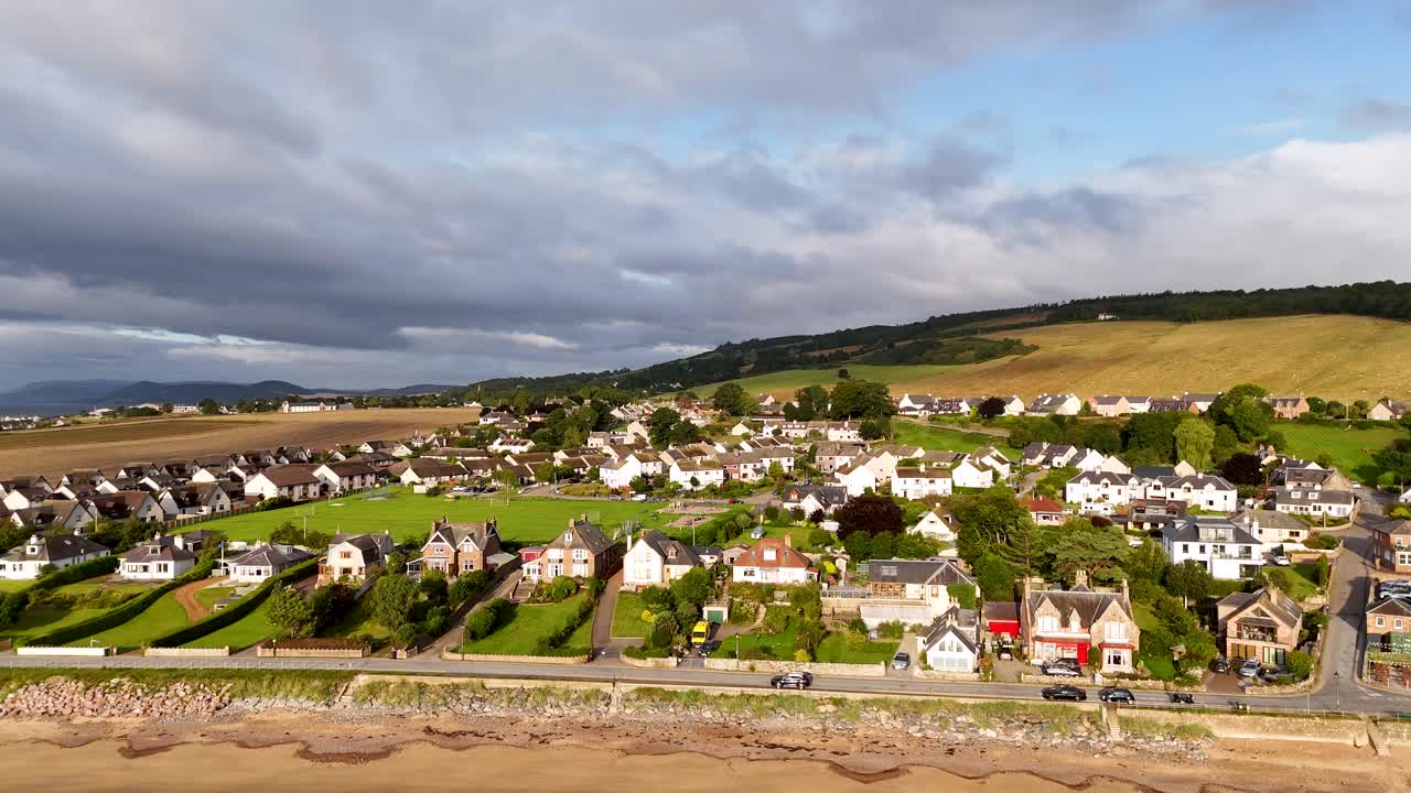 Drone glides toward Rosemarkie shoreline, revealing sandy beach, coastal homes, and rolling Highland hills under soft daylight with dynamic, smooth camera movement