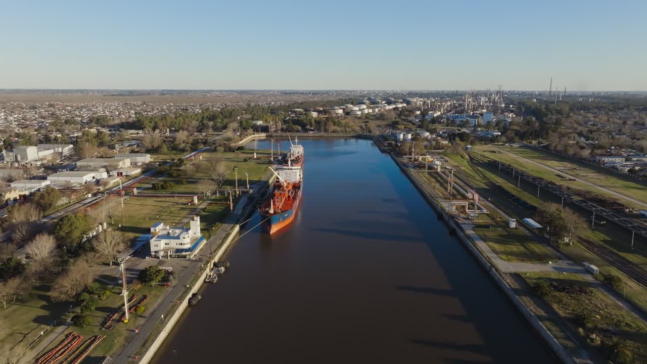 Daytime drone footage capturing a cargo ship at a busy river port with cranes, docks, and surrounding industrial areas