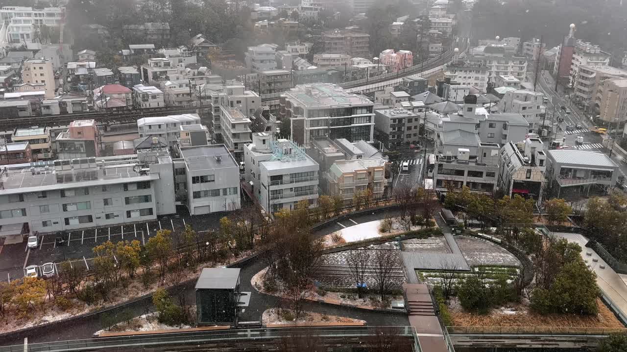 High-rise view of Futakotamagawa on a snowy day, overlooking a small rooftop garden. Snowflakes drift through a grey, overcast sky, capturing a rare and serene winter moment in Tokyo