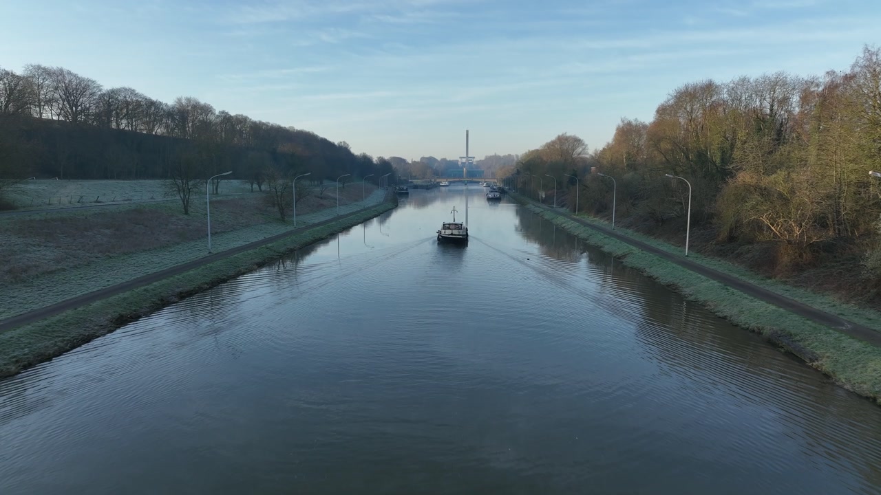 Drone zoom in on a boat cruising the canal near Ronquieres, Belgium, framed by trees and fields under clear blue sky.