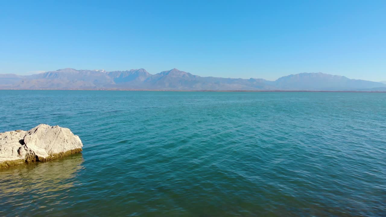 Calm clear water of turquoise lake surface surrounded by high mountains, seen from rocky shore, aerial view