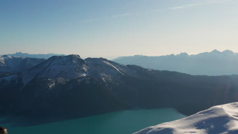 Aerial-dolly-in-past-a-hiker-standing-on-top-of-snowy-mountain-revealing-a-lake