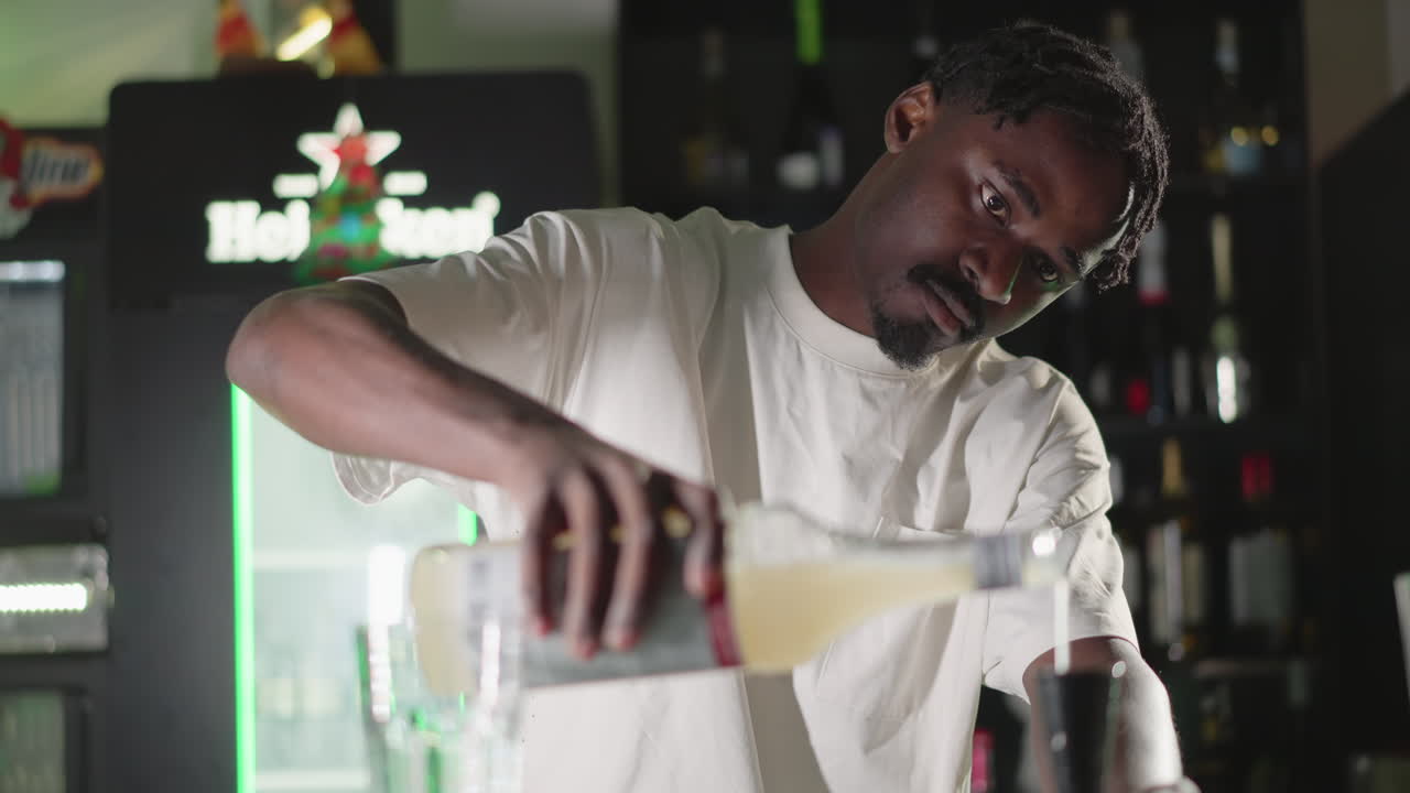 Bartender pouring liquid from bottle into glass, preparing drink at bar counter with focused expression and green neon lighting in background, creating a professional cocktail experience