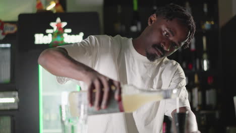 bartender pouring liquid from bottle into glass, preparing drink at bar counter with focused expression and green neon lighting in background, creating a professional cocktail experience