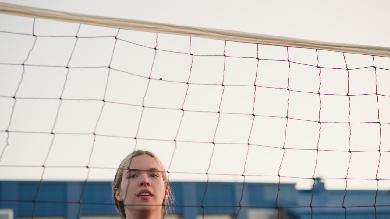 젊은 여성 운동 선수가 점프하고 결단력으로 배구 타격을 준비하는 클로즈업, 그녀의 표정, 배구 네트, 그리고 장면에서 볼 수 있는 배경 건물에 초점을 맞추십시오.