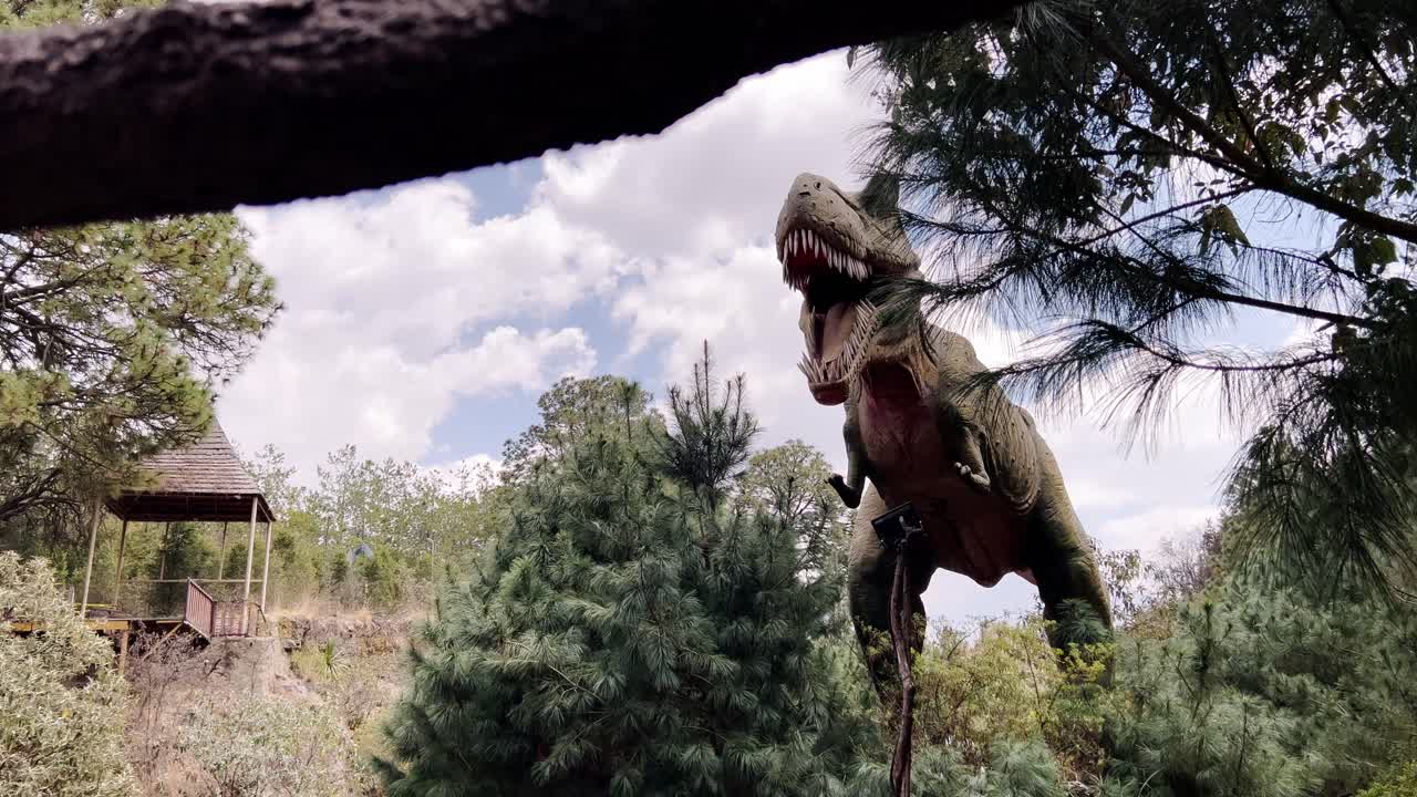Lifelike animatronic T-Rex displayed in wooded outdoor park setting, amid pine trees with a wooden gazebo in the background