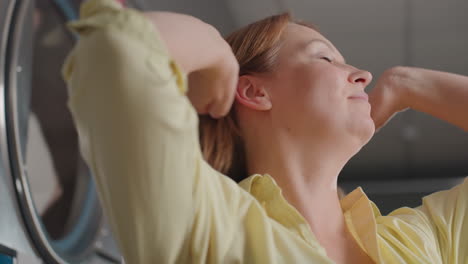 portrait of lady raising hair, arms up, before industrial dryers, clothes rolling behind glass, stainless machines, glowing controls, relaxed smile, laundromat service cleanliness reliability