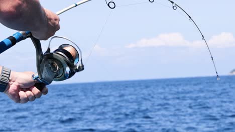 close-up of hands operating a fishing reel against a backdrop of blue ocean and sky.