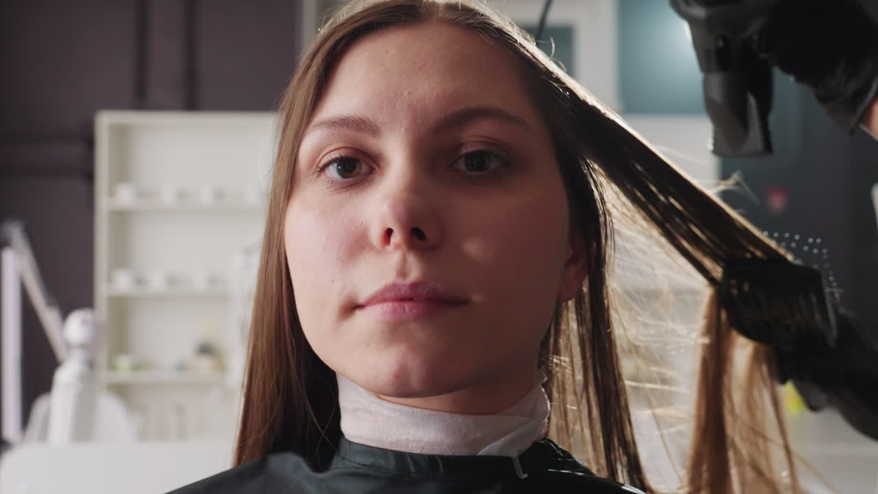 Mujer caucásica sentada en un sillón de peluquería mientras le seccionan el pelo y lo cepillan durante la preparación para el tinte; la estilista lleva guantes negros, iluminación suave de estudio, expresión neutra, capa y espejo visibles, profesional