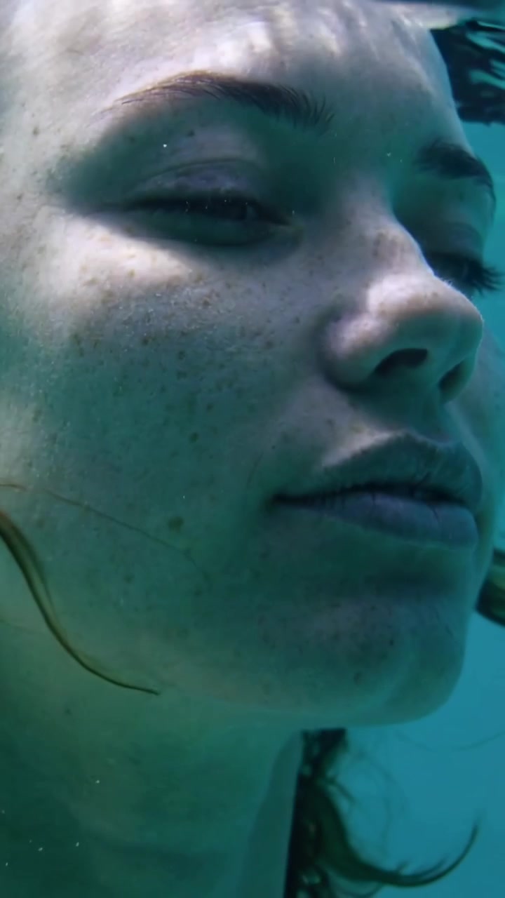 Vídeo vertical: Mulher flutuando de olhos fechados na piscina, com a superfície da água cobrindo a testa, o cabelo e a alça.