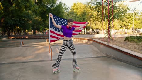 a girl with a short haircut in a purple top and striped pants roller-skates and fits the us flag in a skatepark in summer