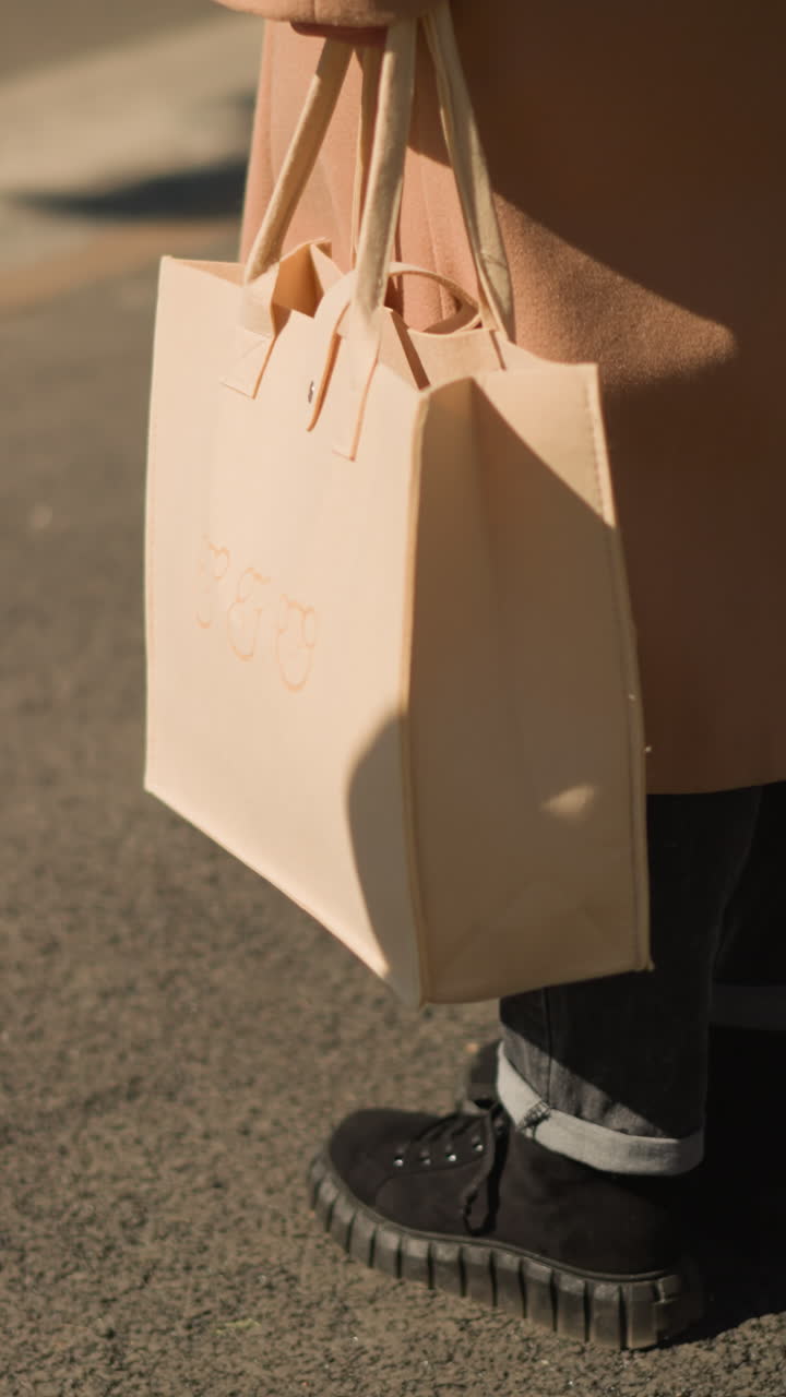 따뜻한 햇살 아래 보도에서 쇼핑객이 들고 있는 베이지색 토트백, 코트 자락과 검은색 부츠 클로즈업, 종이 쇼핑백 손잡이, 은은한 브랜드 엠보싱, 미니멀한 리테일 순간, 가을 거리 분위기, 캐주얼