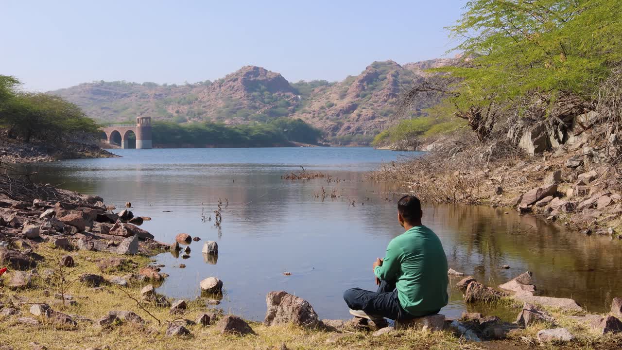 joven aislado sentado en una piedra en la costa del lago prístino por la mañana