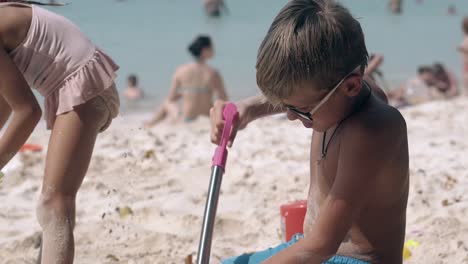 Kleiner-Junge-Mit-Brille-Gräbt-Aktiv-Ein-Loch-Am-Sandstrand