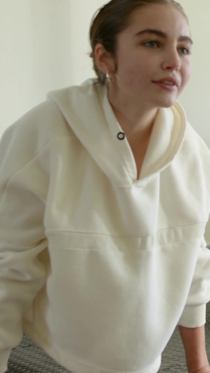 Vertikales Video: Frau in cremefarbenem Kapuzenpullover richtet sich aus einer gebückten Haltung auf einer grauen Matte zu Hause auf und stabilisiert sich.