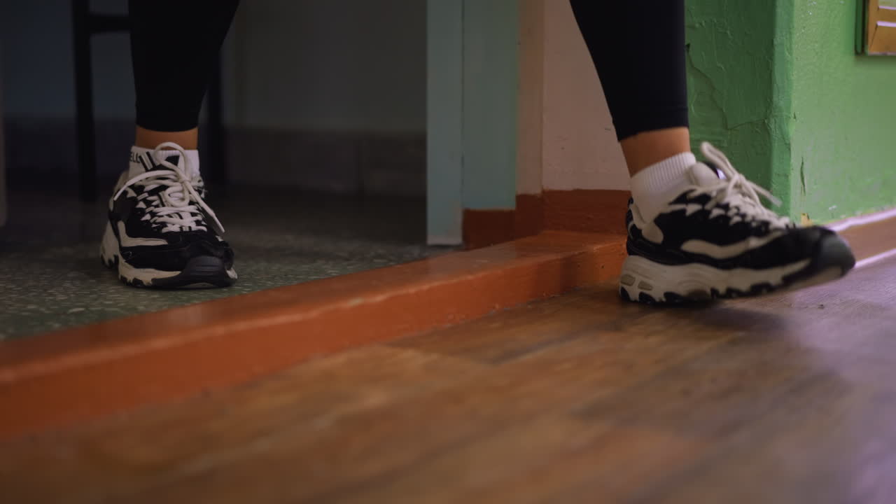 Close view of legs in white and black sneakers stepping out through doorway, casual exit captured with focus on shoes and floor details, indoor background showing textured wall and polished wooden