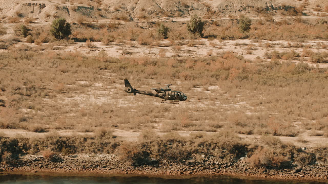 un helicóptero gazela ejecuta hábilmente una maniobra sobre un terreno desolado y seco cerca de un cuerpo de agua. la aeronave muestra su agilidad y precisión en la navegación aérea bajo cielos despejados.