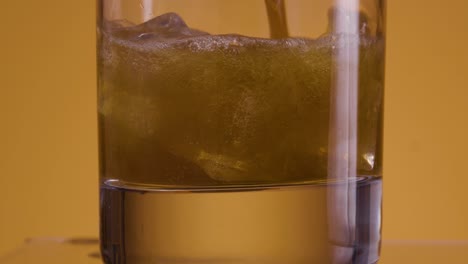 Close-up-of-an-empty-glass-against-golden-yellow-background-with-orange-light-setting-into-which-ice-cubes-fall-and-is-filled-with-cola