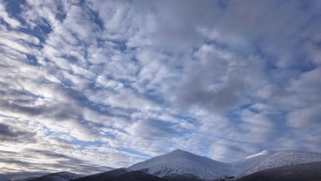 observe the captivating transformation of the sky as clouds drift gracefully over majestic, snow-covered mountains during a tranquil winter day