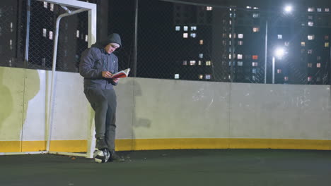 man in hoodie and beanie leaning against metallic goalpost reading book outdoors at night with soccer ball underfoot, scene illuminated by vibrant urban city lights