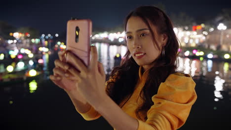 Beautiful-Asian-woman-takes-night-selfie-photo-next-to-river-reflection-in-Hoi-An,-Vietnam