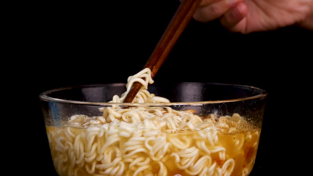 A hand uses chopsticks to stir instant noodles in a clear glass bowl filled with broth, under bright studio lighting against a black background. Close-up, static camera